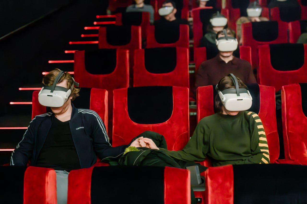 Two viewers in red cinema seats holding hands while wearing VR headsets